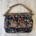 A vintage-style woven tote bag with a dark navy blue background, featuring a detailed tapestry pattern of pinkish-brown rabbits, foxes, and floral motifs, finished with brown leather trim and handles.