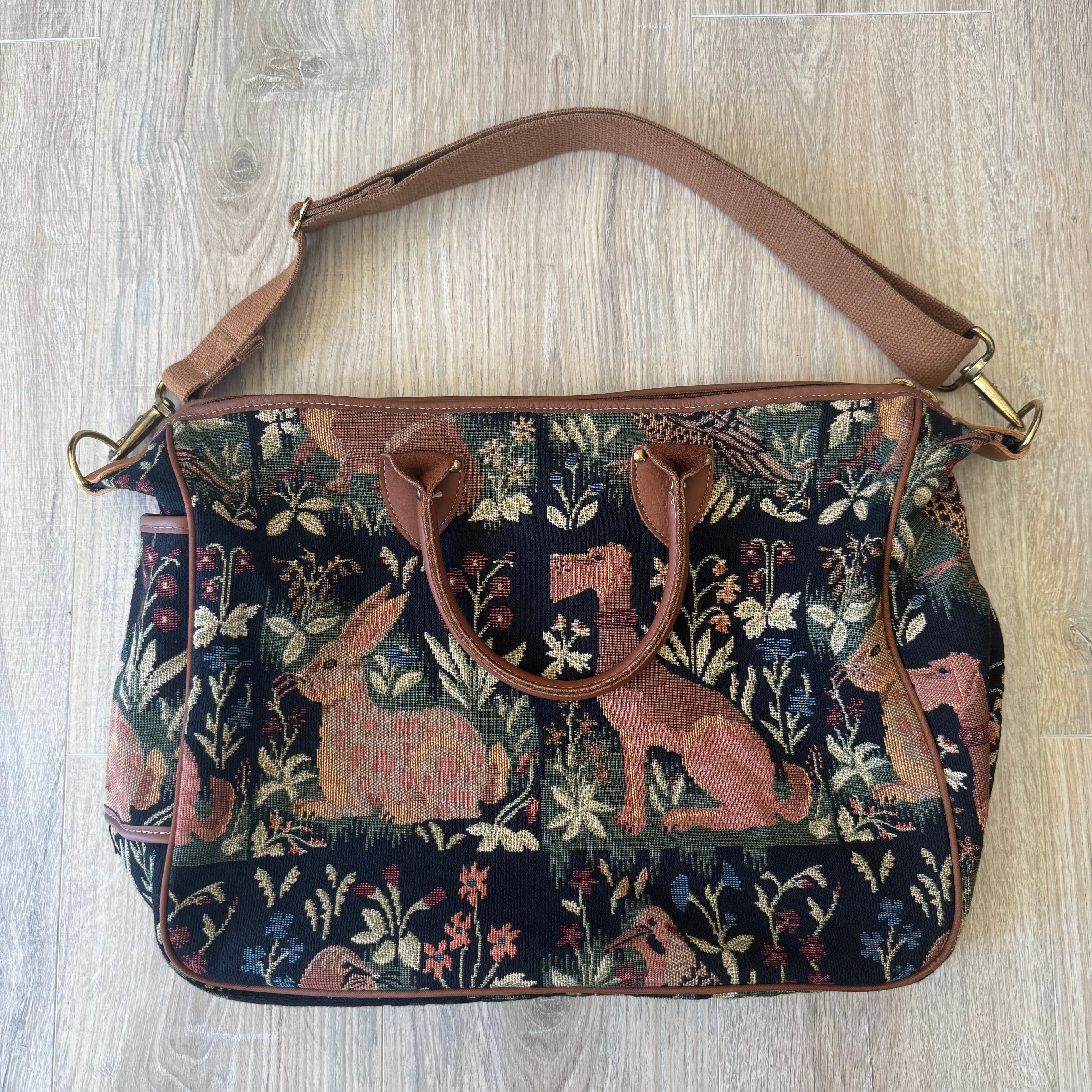 A vintage-style woven tote bag with a dark navy blue background, featuring a detailed tapestry pattern of pinkish-brown rabbits, foxes, and floral motifs, finished with brown leather trim and handles.