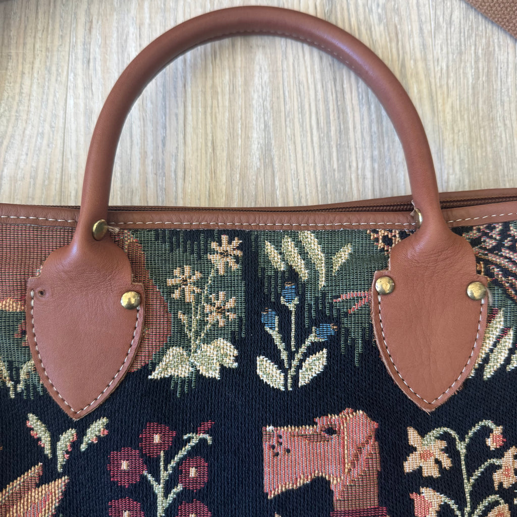 Close-up of a handbag featuring a dark navy blue tapestry with floral and animal motifs, accented with brown leather handles and gold-tone hardware, set against a light wood background.