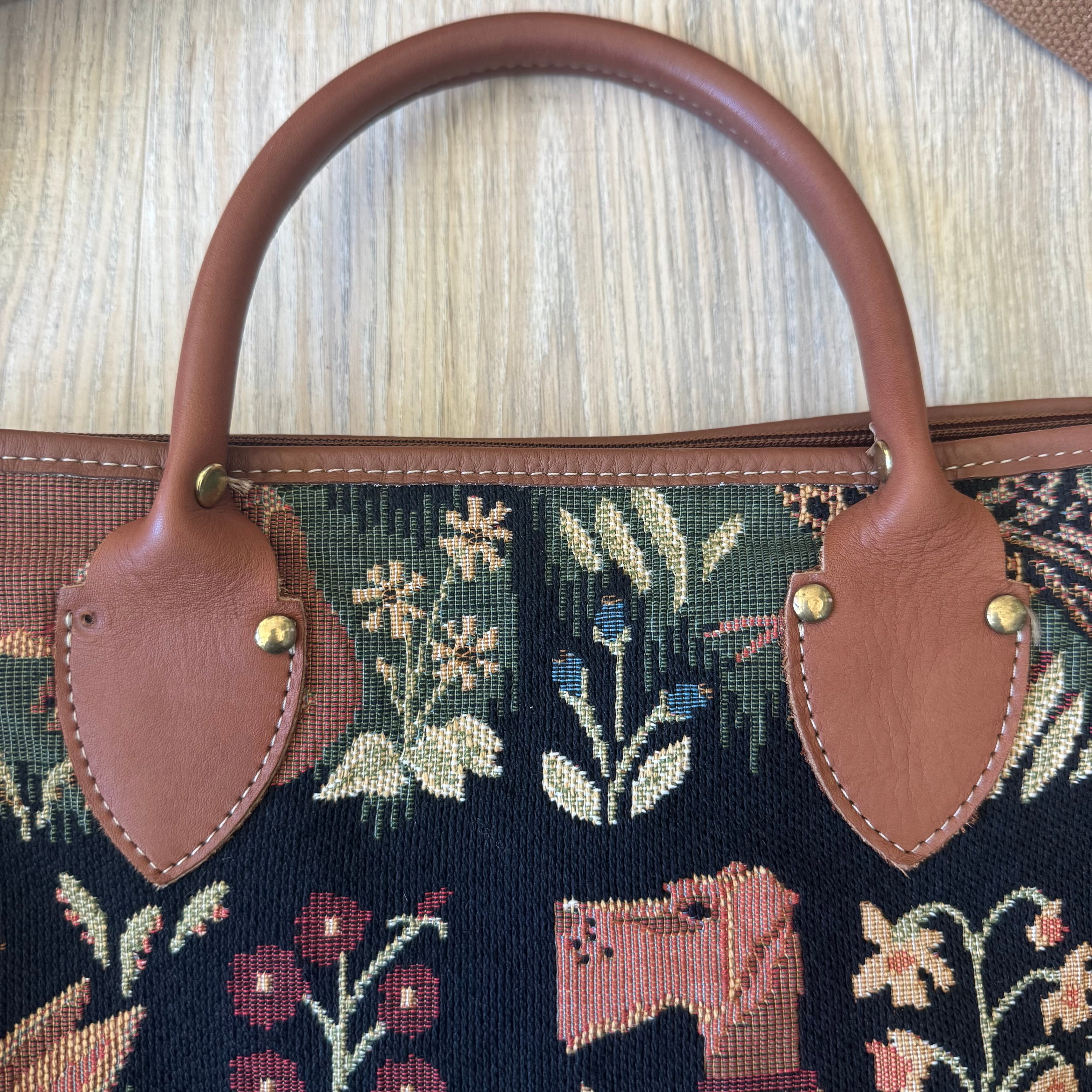 Close-up of a handbag featuring a dark navy blue tapestry with floral and animal motifs, accented with brown leather handles and gold-tone hardware, set against a light wood background.