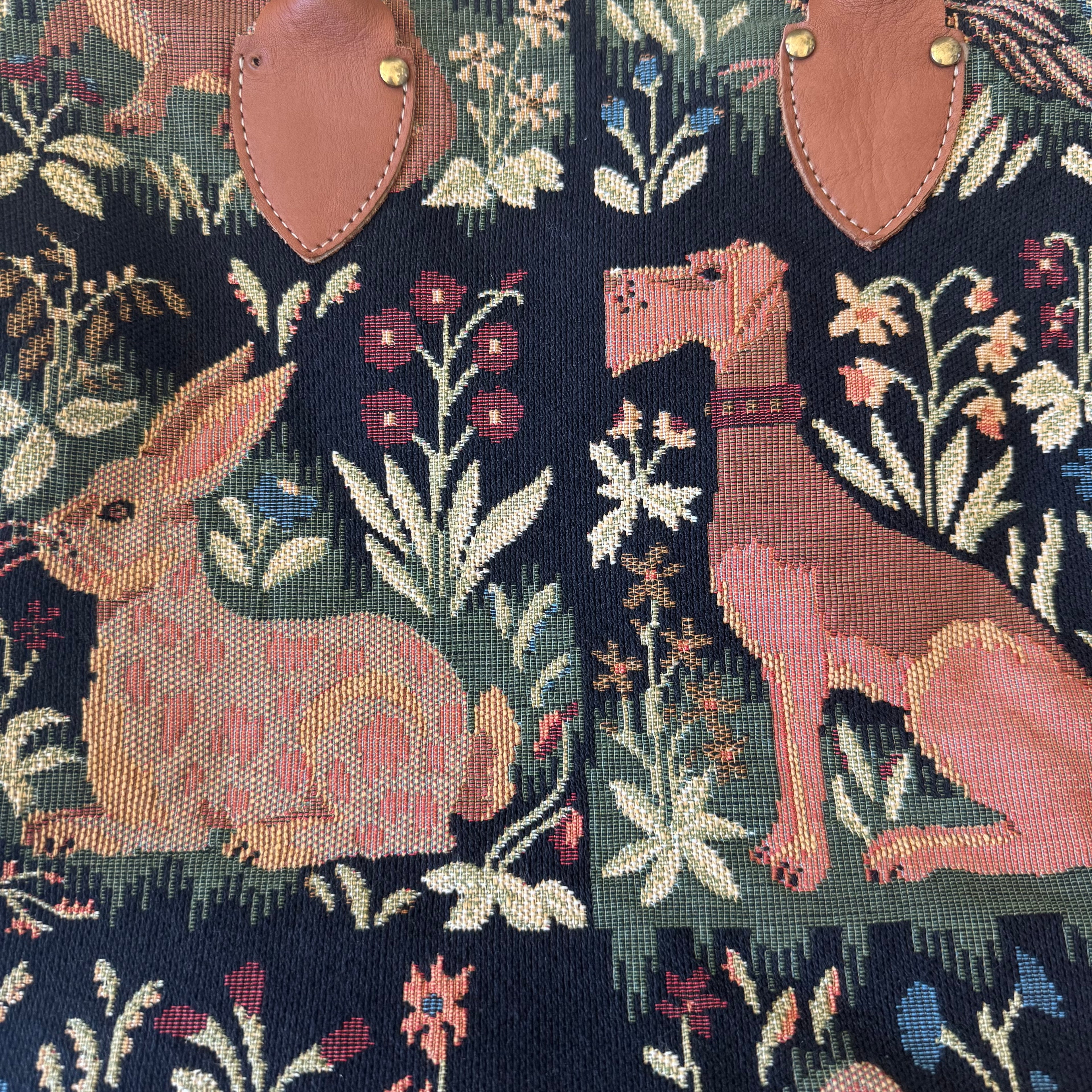 Close-up of a woven textile pattern featuring a pink rabbit, a pink dog, and floral motifs on a dark navy background, with tan leather-like patches and gold-tone metal rivets visible at the top.