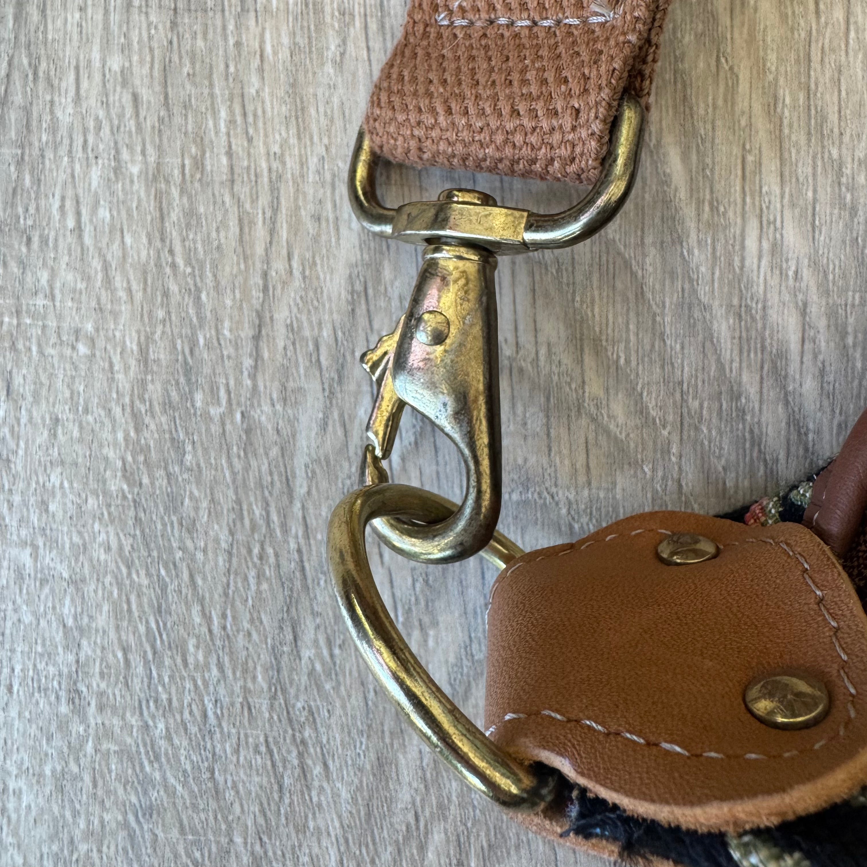 Close-up of a brown woven strap with a brass-tone swivel clasp and metal D-ring, attached to a brown leather loop on a light wood surface.